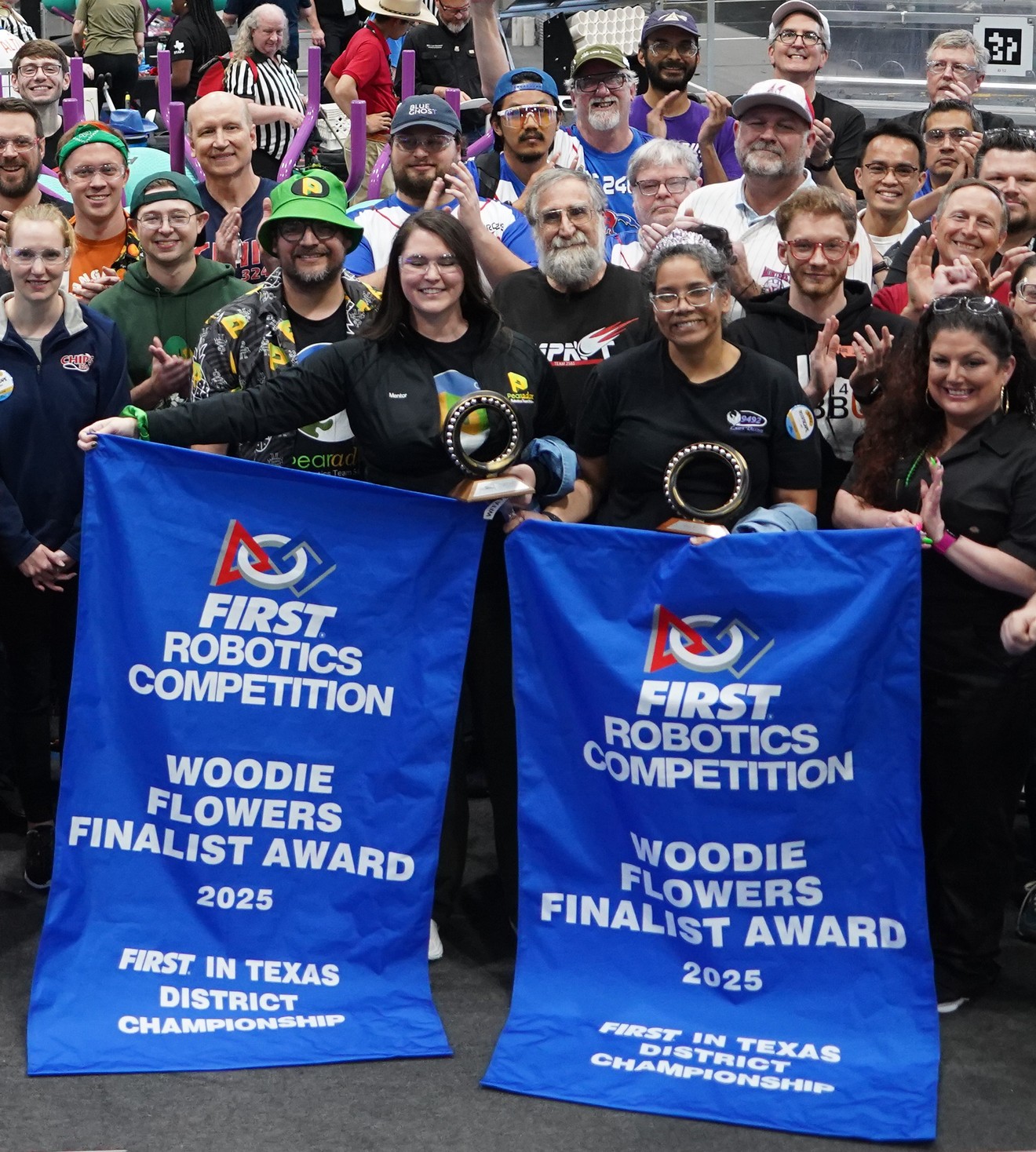 Two WFFA recipients holding their blue Woodie Flowers Finalist Award banners at the 2025 FIRST in Texas District Championship, surrounded by cheering crowd