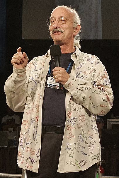 Dr. Woodie Flowers speaking on stage holding a microphone and wearing a signature signed shirt