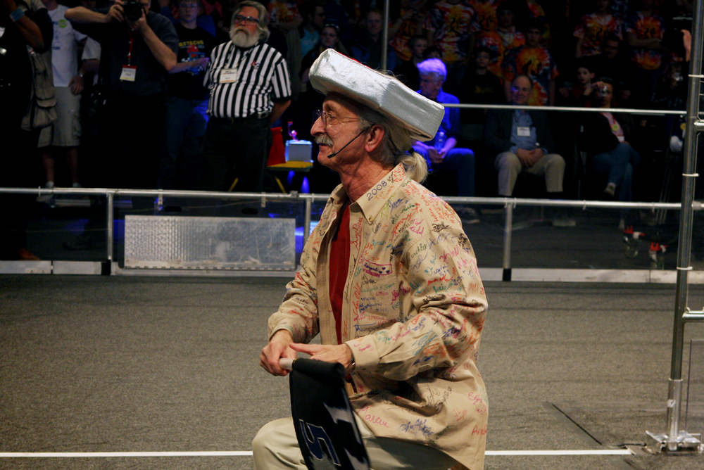 Dr. Woodie Flowers wearing a signature signed shirt and a large silver hat on a competition field