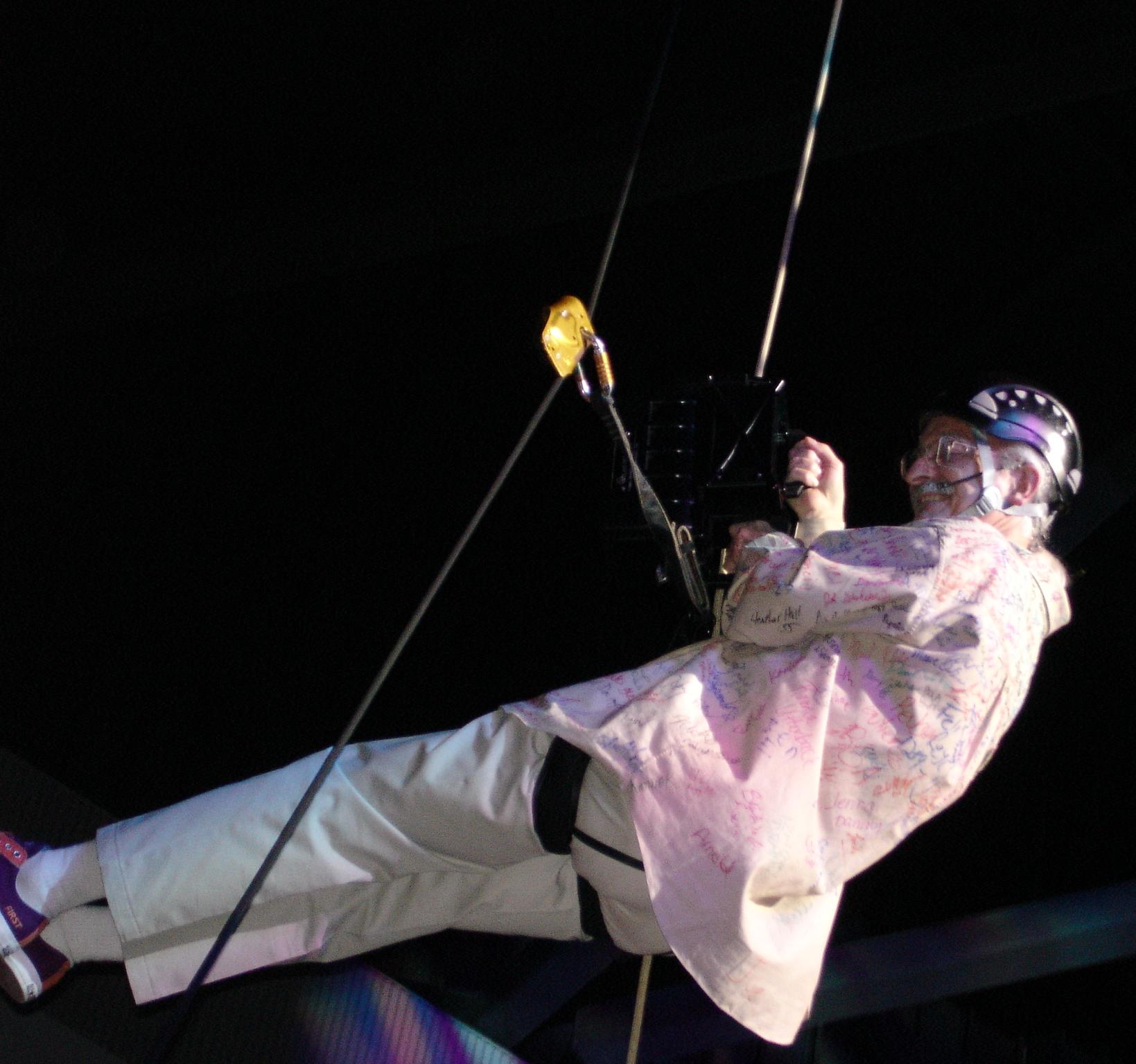 Dr. Woodie Flowers rappelling down in a harness and helmet wearing a signature signed shirt