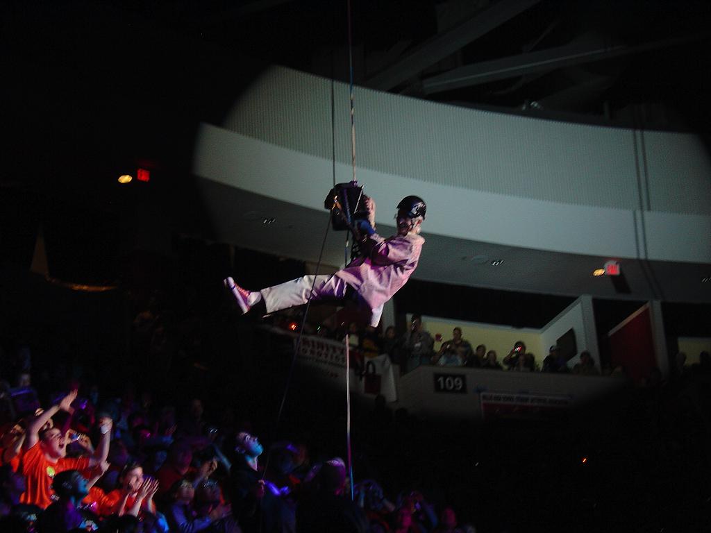 Wide shot of Dr. Woodie Flowers rappelling down from the arena ceiling into the cheering crowd at a FIRST event