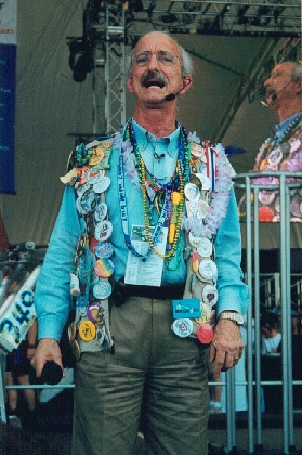 Dr. Woodie Flowers standing on stage wearing a vest covered in FIRST buttons, pins, and beads