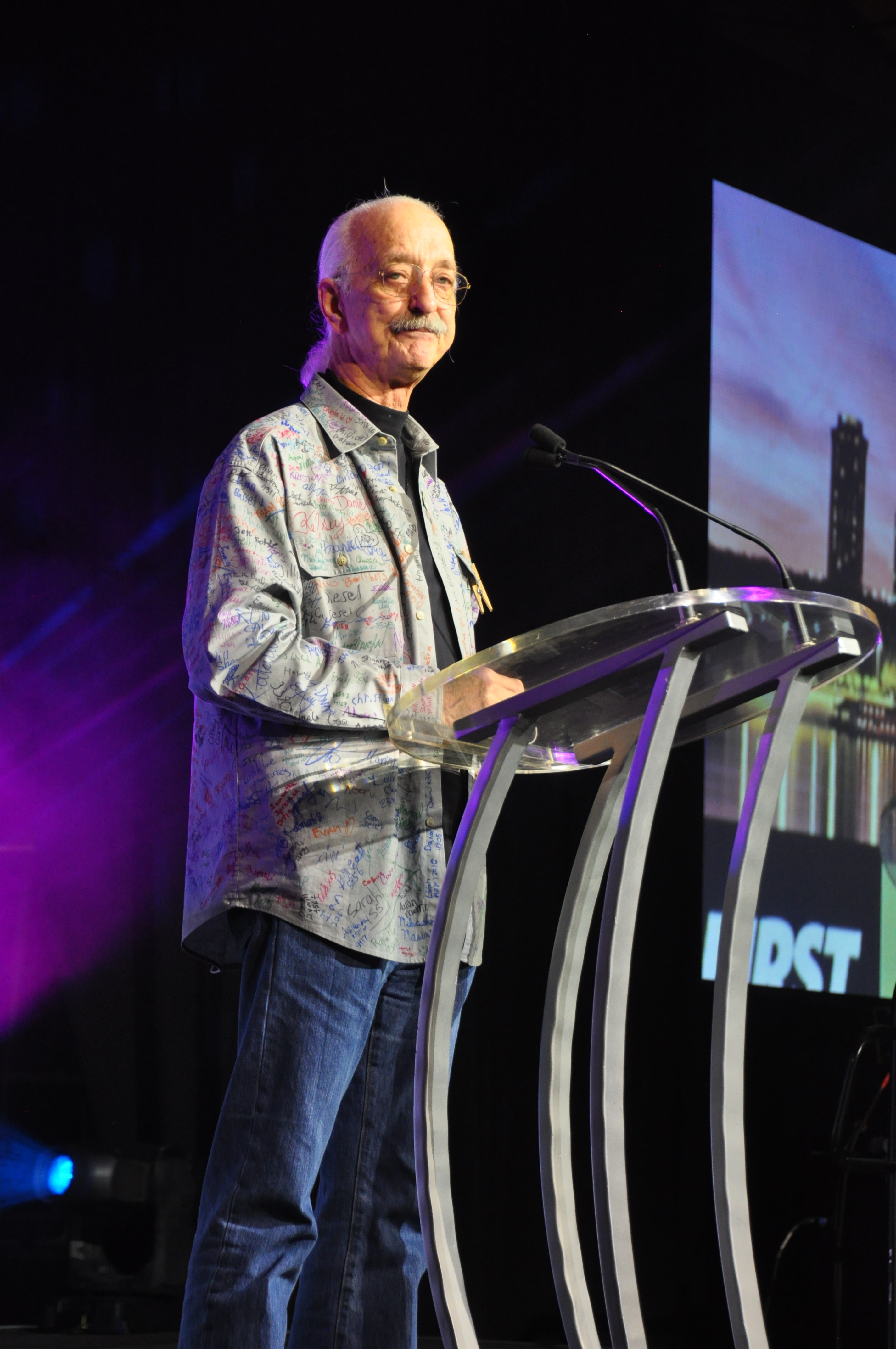 Dr. Woodie Flowers speaking at the 2017 FIRST Championship at a glass lectern wearing a signed shirt