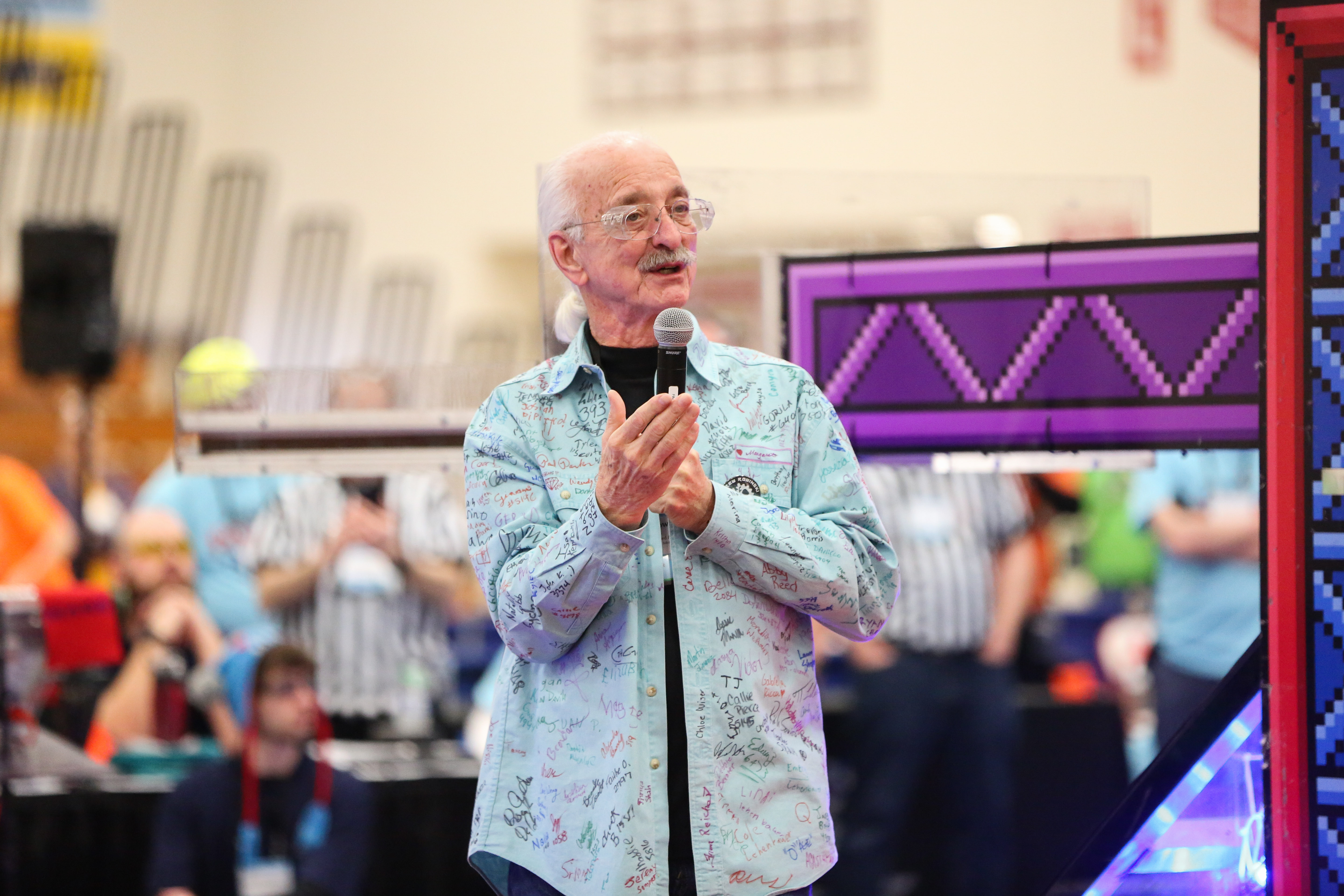 Dr. Woodie Flowers speaking into a microphone at the 2018 FRC Livonia district event wearing a signed shirt